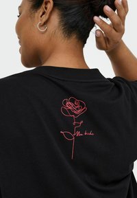Zwarte katoenen t-shirt met een rode lijntekening van een roos en de tekst "Ma biche" op de achterkant. Korte mouwen en een ronde hals.