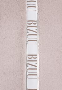 Sztuczne futro w jasnym beżu o miękkiej teksturze, z białą wstążką z brązowym napisem "BIZUU" wzdłuż krawędzi.