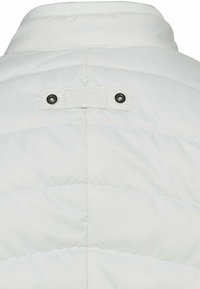 Dos de veste matelassée blanche avec une petite étiquette en tissu cousue et deux œillets métalliques noirs près du col.