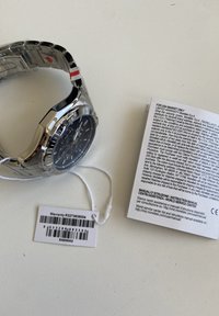 Sølv metalur ur med sort urskive, chronograf funktioner og et armbånd i rustfrit stål. Inkluderer garanti-tag og brugsanvisning.