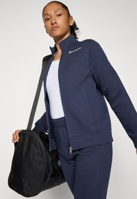 Femme portant un survêtement Champion bleu marine et un haut blanc, tenant un sac de sport noir sur l'épaule devant un fond neutre.
