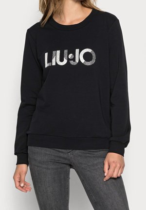 Kobieta ubrana w czarną bluzę z srebrzystym, błyszczącym logo "LIU JO" na piersi oraz ciemnoszare dżinsy, stojąca na jednolitym tle.