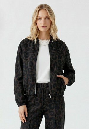 Femme blonde portant une veste noire à imprimé léopard et un pantalon assorti avec une chemise blanche, debout avec une main dans la poche de la veste.