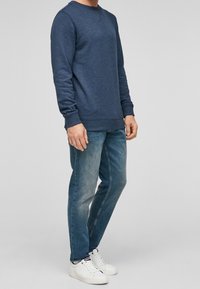 Sudadera azul de cuello redondo en tejido texturizado, combinada con vaqueros de tono azul claro y zapatillas blancas. Puños y dobladillo acanalados visibles.