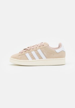 Beige suède Adidas Campus sneaker met witte strepen en hielstuk, veterontwerp en een gebroken witte rubberen zool, gepresenteerd in profiel.