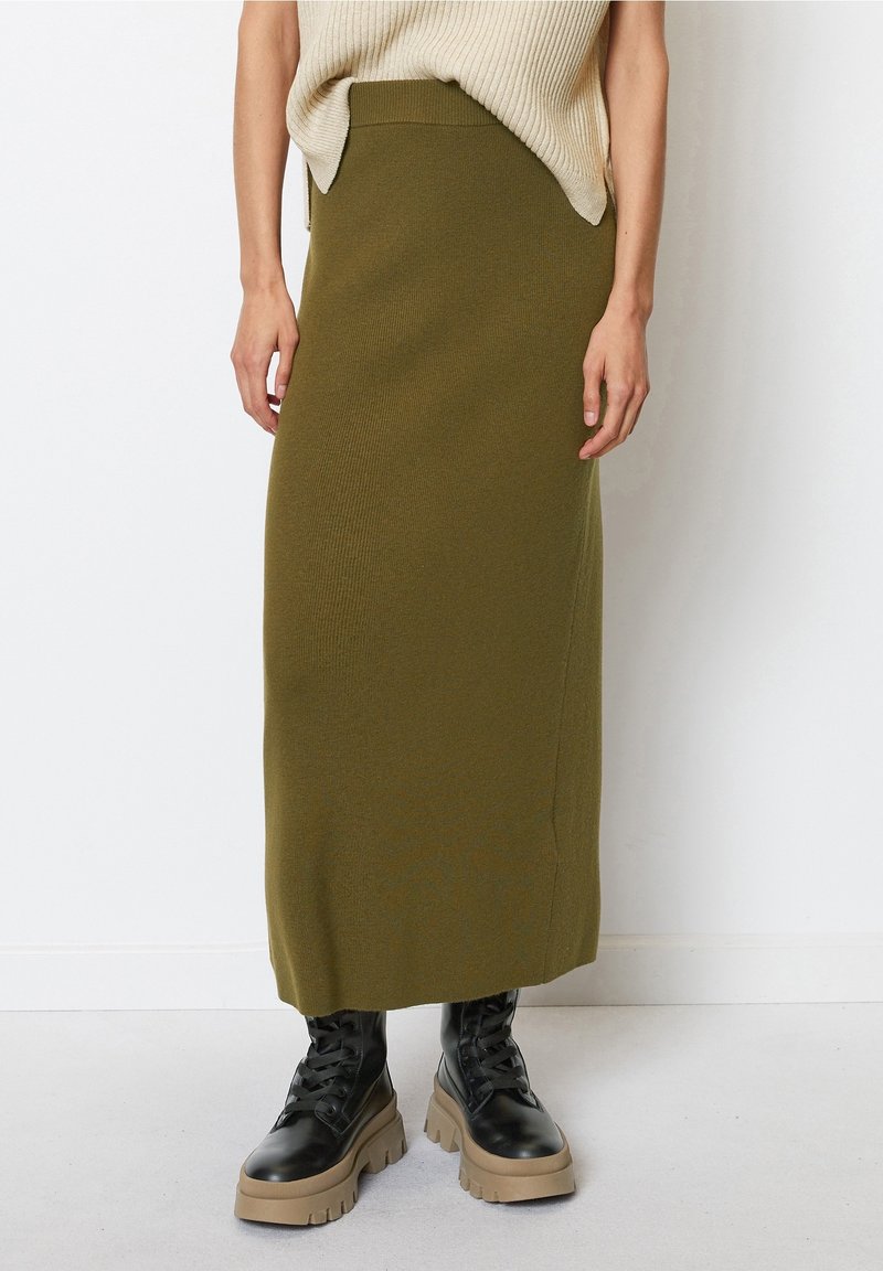 Femme portant une jupe midi tricotée vert olive, un haut beige sans manches et des bottes noires à plateforme épaisses, debout contre un mur blanc.