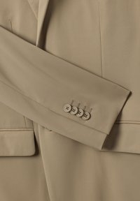 Maniche di giacca beige con quattro bottoni chiari, rifiniture cucite e una texture di tessuto liscia. Presenta un design su misura e linee affilate.