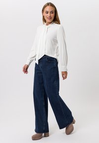 Witte blouse met een nekdas, lange mouwen. Gepaard met donkerblauwe wijde jeans en lichtbruine plateauschoenen. Effen achtergrond.