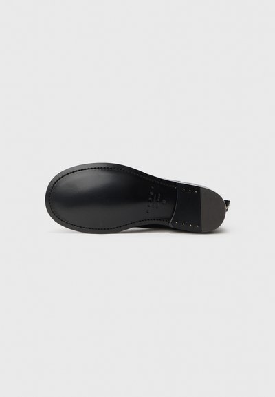 Sole di scarpa in pelle nera con una finitura liscia, punta arrotondata e cuciture visibili. Presenta un tacco basso e borchie metalliche lungo il bordo.