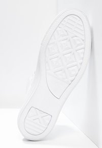 Vit sneakersula med geometriska mönster, ställd upprätt mot en vit hörnvägg och golv.