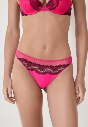 Femme portant de la lingerie rose vif avec une finition en dentelle violette foncée, montrant le milieu du corps et le haut des cuisses sur un fond blanc uni.