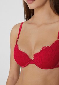 Červená podprsenka s krajkovým překrytím, hladkými okraji a nastavitelnými ramínky. Texturovaný pás a květinové vzory zdobí košíčky.