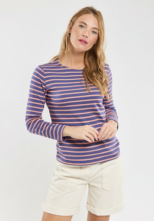 Jeune femme aux longs cheveux blonds portant un t-shirt à manches longues rayé bleu marine et corail et un short blanc retroussé sur un fond uni.