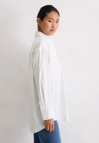 Junge Frau mit dunklen Haaren, die nach hinten gebunden sind, trägt ein lockeres weißes Hemd und blaue Jeans und steht seitlich gegen eine weiße Wand.