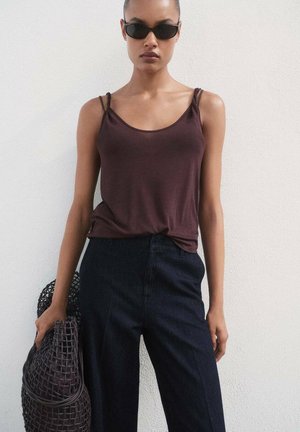 Bordeaux kleurige tanktop met dunne spaghettibandjes, gecombineerd met donkere denim hoge taille broeken. Model houdt een gestructureerde geweven tas vast.
