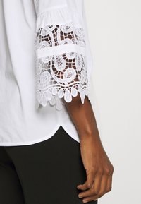 Blusa blanca con intrincados bordados de encaje en las mangas, con un dobladillo redondeado. Combinada con pantalones oscuros ajustados.
