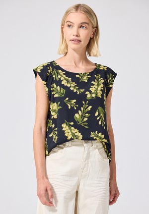Femme aux cheveux blonds courts porte un haut noir sans manches à imprimé floral jaune et un pantalon blanc, regardant légèrement sur le côté.
