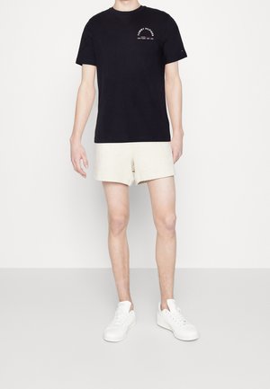 Musta puuvillainen t-paita rintalogo edessä, yhdistetty vaaleanbeigeihin shortseihin. Mallilla on valkoiset lenkkarit. Yksinkertainen, rento asu siistillä leikkauksella.