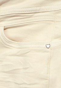 Närbild av en beige denimficka med en liten vit hjärtformad nitar i övre högra hörnet av fickan.