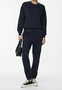 Marinblå sweatshirt och matchande joggers i mjukt material, med dragsko i midjan, muddar vid anklarna och svarta sneakers. Svart handväska.