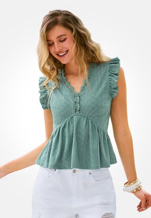 Lächelnde Frau, die ein ärmelloses, türkisfarbenes Spitzenoberteil mit Rüschen und weiße, ausgefranste Shorts trägt, dazu Armbänder als Accessoires.