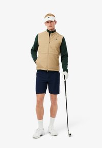 Chaleco acolchado beige sobre una camisa verde de manga larga, combinado con pantalones cortos azul marino. Visera blanca, guante y palo de golf completan el conjunto.