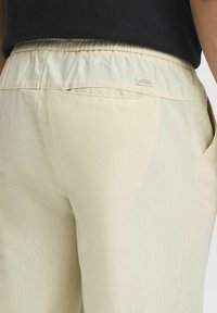 Lys beige bukser med elastisk talje, sidelommer og et lille mærke, der står "Dedicated to Denim Perfection." Teksturert stof.