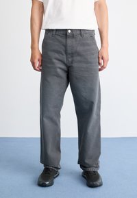 Persona che indossa pantaloni grigi larghi, sneakers nere e una maglietta bianca a maniche corte, in piedi su un pavimento blu contro un muro bianco.