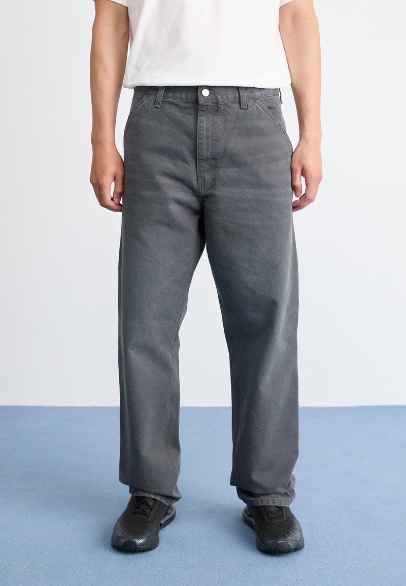 Persona che indossa pantaloni grigi larghi, sneakers nere e una maglietta bianca a maniche corte, in piedi su un pavimento blu contro un muro bianco.