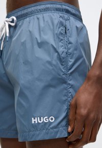 Jasnoniebieskie spodenki kąpielowe wykonane z lekkiego materiału, posiadają elastyczną talię z troczkiem oraz biały napis „HUGO” na dole.