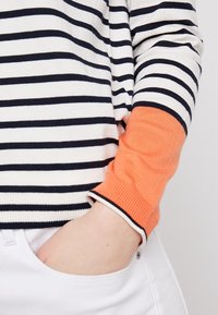 Maglione a righe con strisce orizzontali in blu navy e crema, con un polsino a coste arancione. Il materiale sembra morbido; l'orlo ha un bordo bianco.