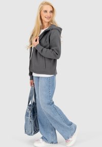 Felpa grigia con zip e cordoncini, abbinata a una camicia bianca e jeans in denim blu a gamba larga. Il modello tiene una borsa grigia.