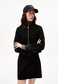 Sort fløjljakke med lynlås foran og lomme, parret med en matchende sort fløjlskørt. Modellen bærer en sort cap.