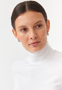 Valge soonikkoes turtleneck-särk kõrge kraega, koos hõbedaste r�õngaskõrvarõngastega. Ühtlane taust toob esile rõiva tekstuuri.