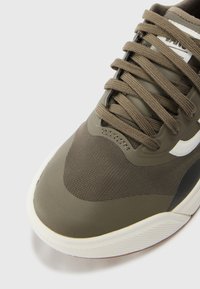 Baskets vert olive avec une tige en mesh, des superpositions texturées et des lacets ronds. Accents blancs sur le logo et la semelle. Design sportif classique.