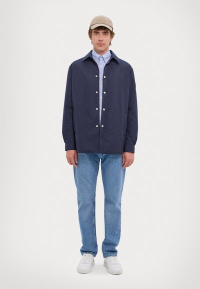 Mørkeblå, afslappet pasform jakke med spids krave, der har sølvknapper nede foran. Lys blå skjorte med krave og lyse jeans.