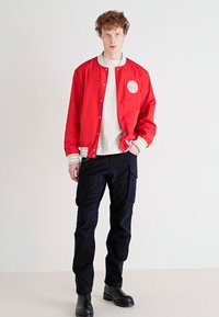 Red bomber jacket with white accents, featuring a logo patch. Paired with black cargo pants and black boots, set against a neutral background.