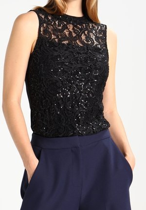 Femme portant un haut en dentelle noire sans manches avec des sequins et un pantalon taille haute bleu marine, mains dans les poches sur fond uni.