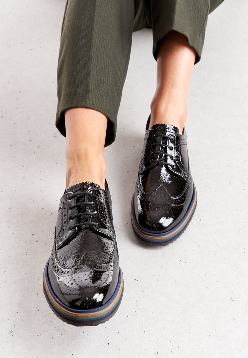 Chaussures à lacets en cuir verni noir avec détails brogue et semelle à rayures arc-en-ciel, présentant une finition texturée et des bords festonnés.
