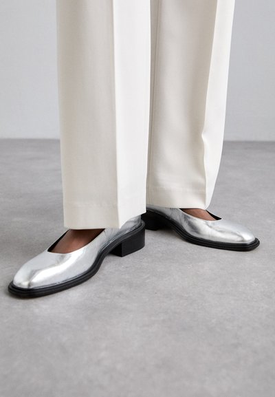 Hõbedased nahkkingad madala musta ploki kontsaga ja mandlilaadse ninaga, sobitatud heledate pükstega. Sile, peegeldav tekstuur.