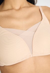 Lys rosa bh med en strukturert bølgedesign. Har et kryss-design i gjennomsiktig materiale og justerbare stropper. Glatt finish og myk tekstur.