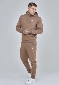Bruine hoodie en bijpassende joggers gemaakt van zachte stof, met een klein wit logo op de borst en een strakke pasvorm rond de manchetten en enkels.