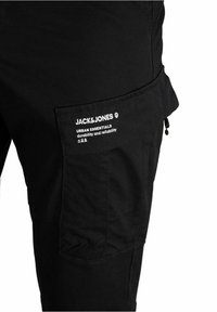 Svarta cargo-byxor med en sidoficka som har texten "JACK & JONES Urban Essentials hållbarhet och pålitlighet" samt en liten dragkedja.