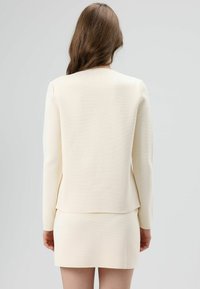 Ensemble blazer et jupe crème clair, tissu texturé, coupe ajustée, manches longues, détail de couture au dos, silhouette élancée, esthétique minimaliste.