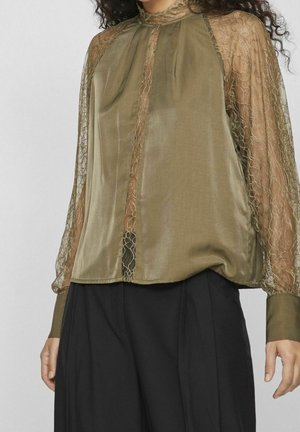 Vrouw draagt een olijfgroene doorschijnende kanten blouse met lange mouwen en hoge hals en zwarte broek met wijde pijpen.