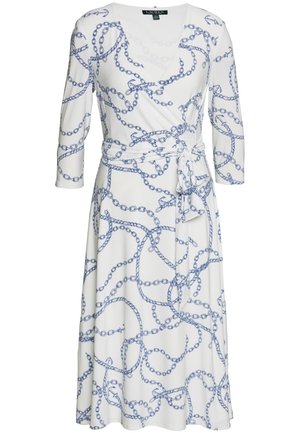 Mujer con un vestido cruzado blanco con estampados de cadenas azules, zapatillas blancas y un bolso blanco de hombro sobre un fondo liso.