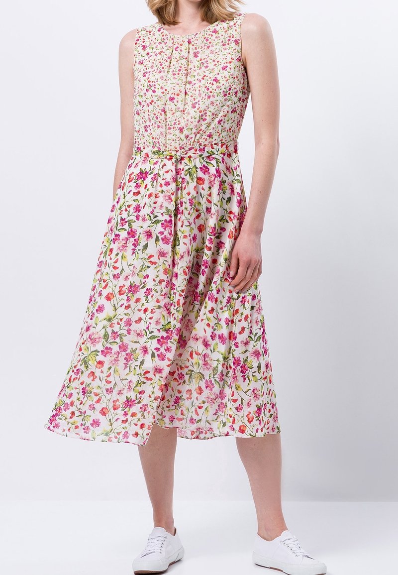 Robe midi à fleurs blanche avec des motifs roses et verts, sans manches, avec un corsage plissé et une jupe fluide. Associée à des baskets blanches.