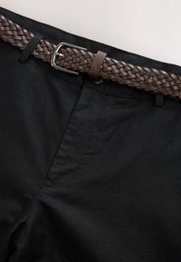 Ceinture en cuir tressé marron passée dans les passants d’un pantalon noir, sécurisée par une boucle argentée.