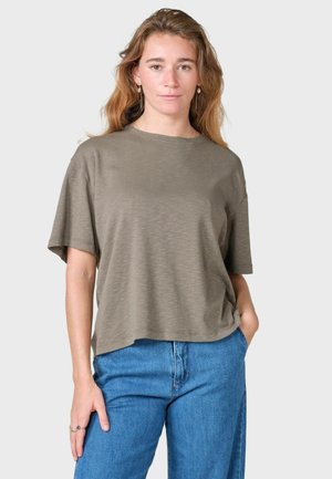 Jeune femme aux cheveux ondulés porte une chemise ample à manches courtes vert olive et un jean bleu, debout avec une main dans la poche.