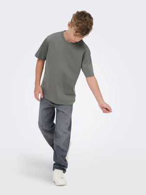 Boy in gray-green T-shirt, dark jeans, and white sneakers standing with head down and right arm extended sideways against plain background.